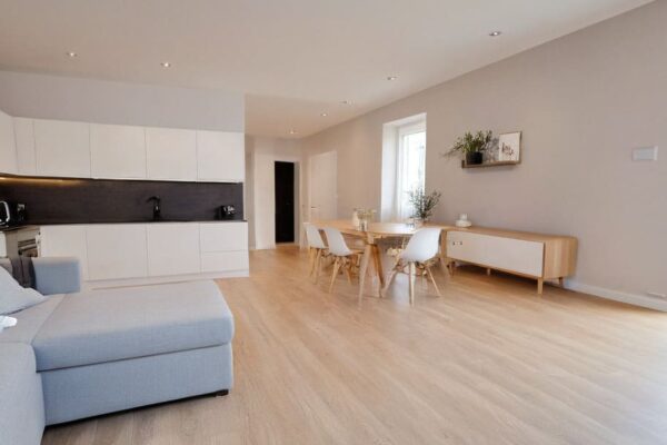 Nowoczesne, wykończone pod klucz wnętrze prezentujące jasny salon z aneksem kuchennym, przedstawione jako inspiracja dla osób poszukujących mieszkania na sprzedaż.